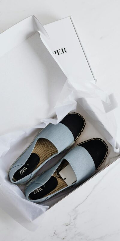 Top view of fashionable espadrilles in an open white cardboard box on a marble surface.