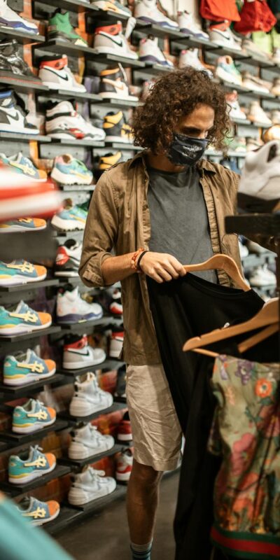 Young man in a sneaker and clothing store wearing a mask, browsing through shirts amid pandemic.
